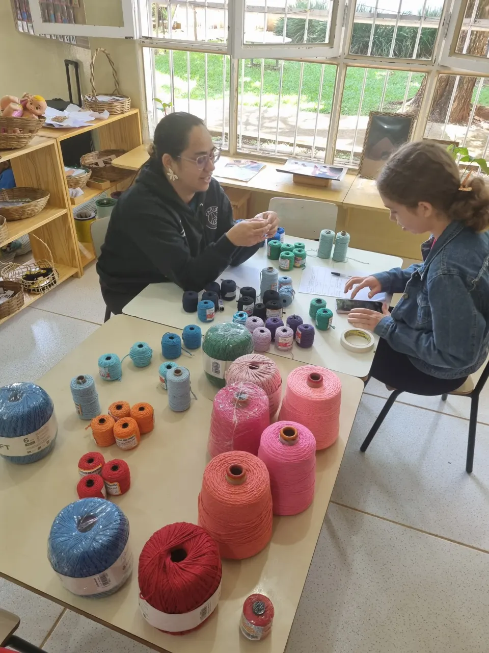 Duas educadoras trabalhando com novelos e cones de linha coloridos sobre a mesa