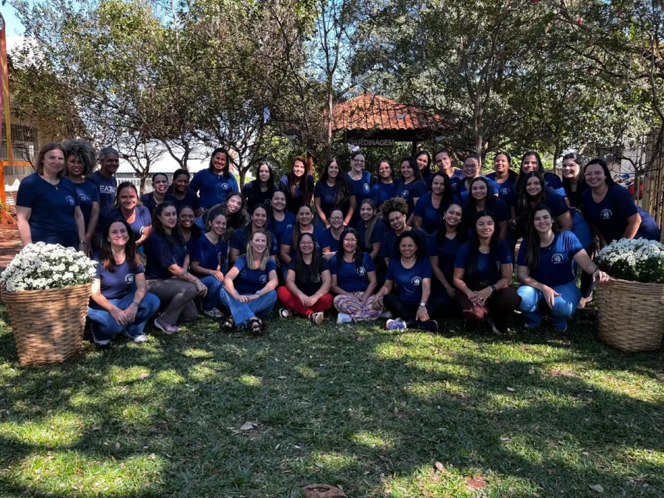 Equipe da creche reunida, uniformizada, no gramado do jardim, entre dois cestos de flores brancas